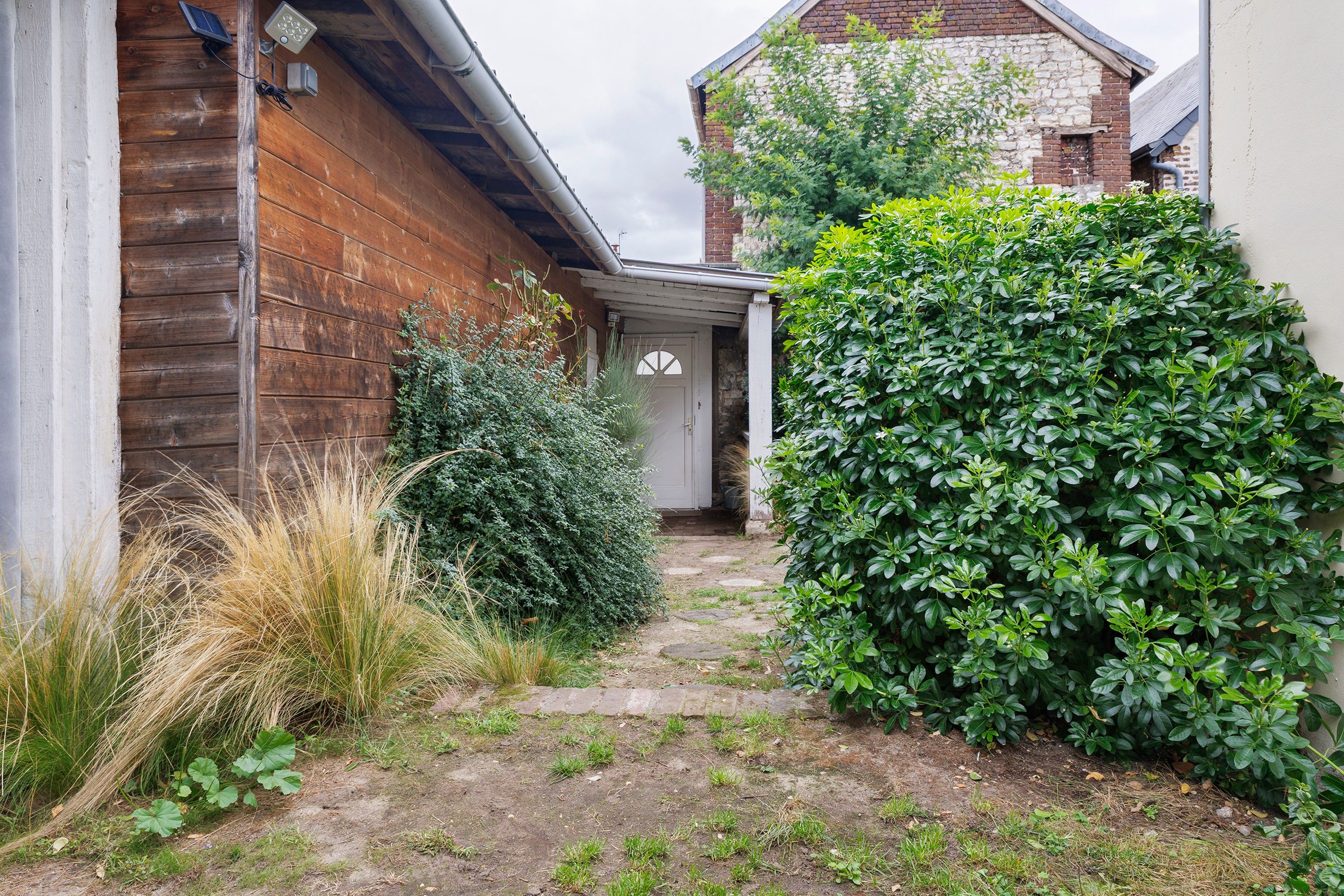 Un étroit sentier menant à une porte, bordé d’un mur en bois et d’un buisson.