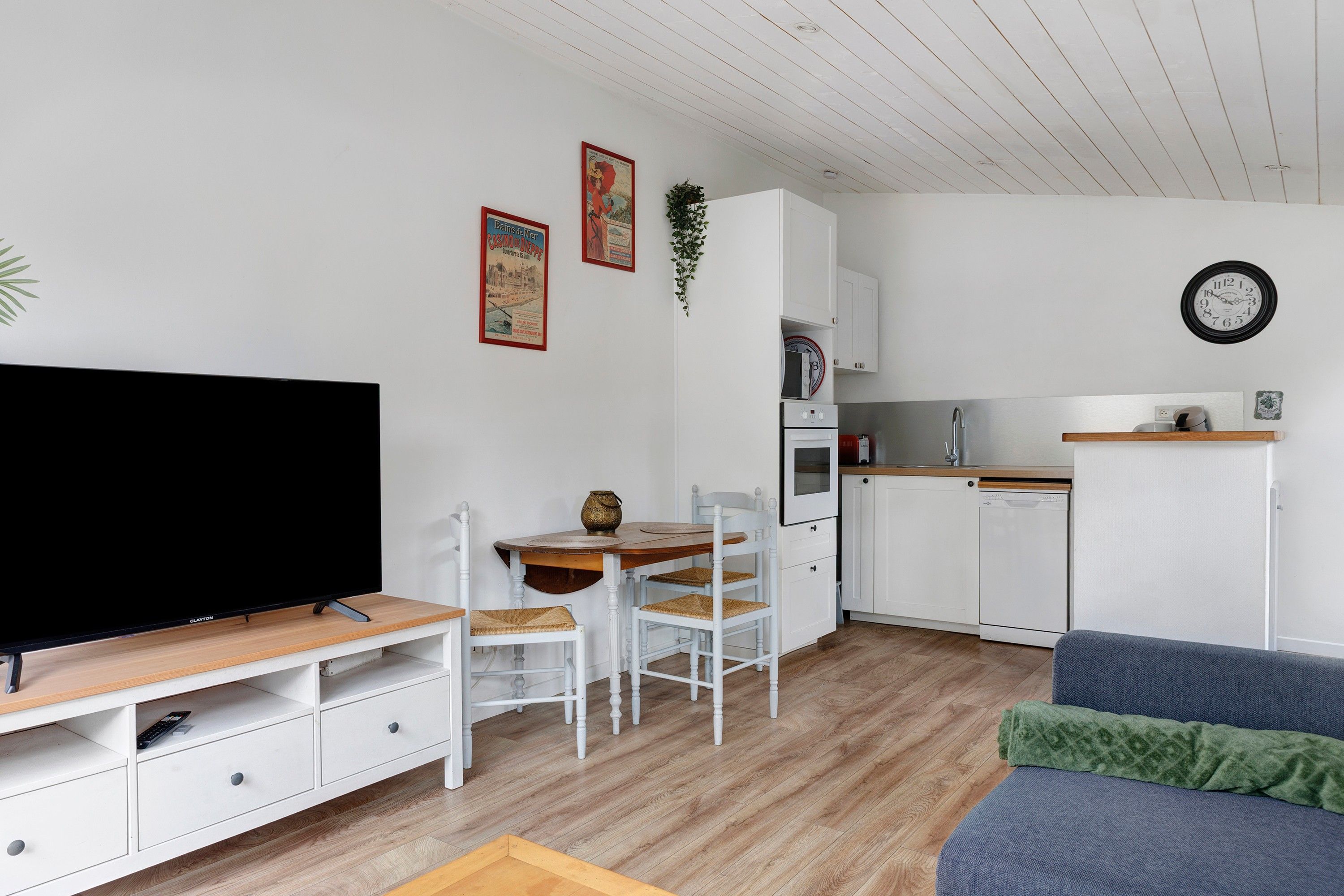 Een moderne woonkamer met een flatscreen televisie, een eettafel met stoelen, een keuken met kasten en apparatuur, en een klok aan de muur.