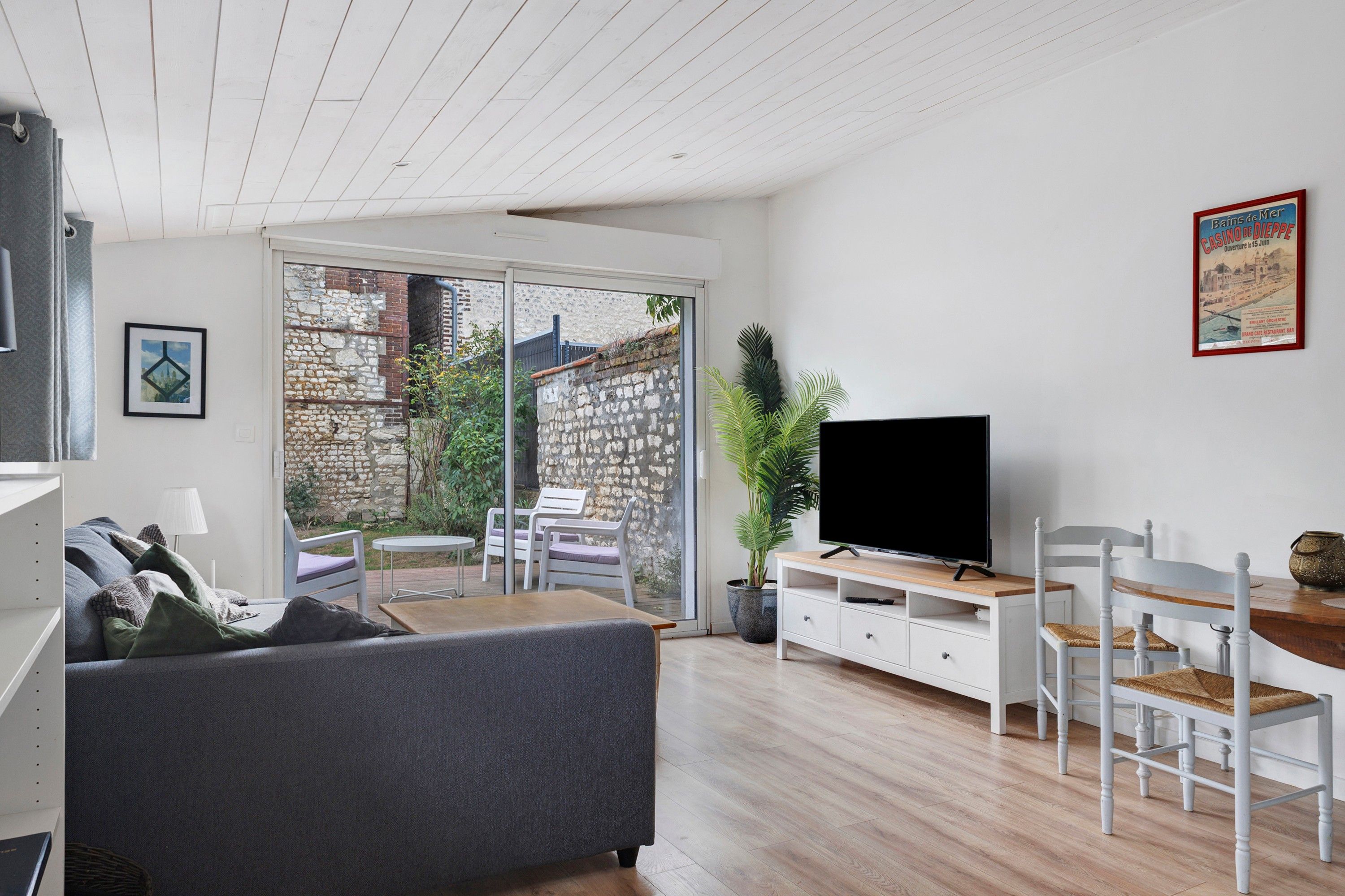 Un salon moderne avec un canapé gris, un téléviseur posé sur un meuble en bois, un coin repas avec une table et des chaises, et une grande fenêtre donnant sur un mur de pierre et de la verdure.
