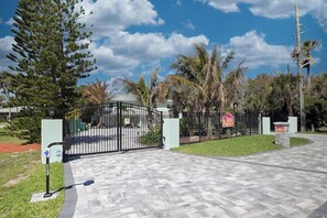 A gated entrance with a paved driveway, palm trees, and a clear sky. - Oasis Incredible River Front/Ocean Access w/Dock (Melbourne Beach)