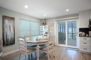 A dining area with a round table, four chairs, a kitchen with cabinets, a clock, and a view through a glass door. - Oasis Incredible River Front/Ocean Access w/Dock (Melbourne Beach)
