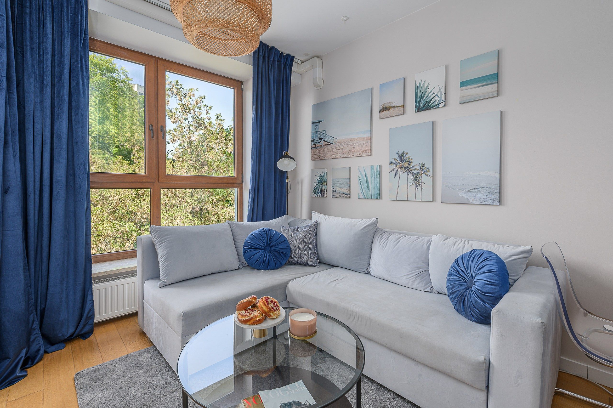 A modern living room with a grey sofa, blue cushions, a glass coffee table, and beach-themed artwork on the wall.