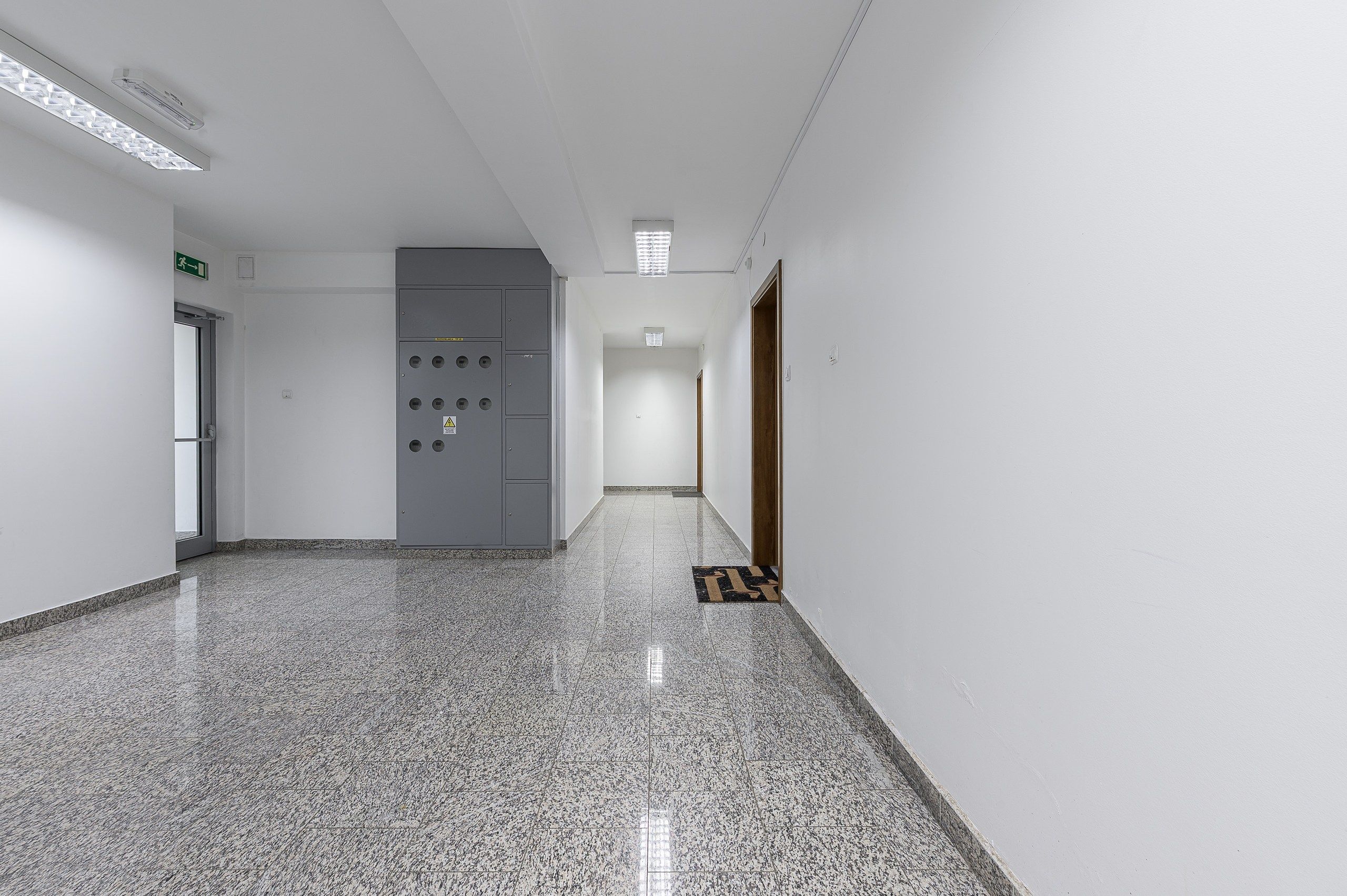 A hallway with a tiled floor, a grey electrical panel, and a door with a wooden frame.