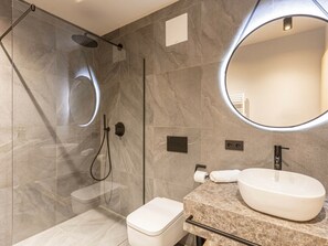 Un baño moderno con ducha envuelta en vidrio, un espejo redondo, un lavamanos blanco y un inodoro.