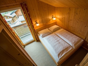A wooden cabin room with a bed, two wall-mounted lights, and a view of a wooden deck and buildings outside. - Cabin in Annaberg With Swimming Pool (Annaberg-Lungoetz)