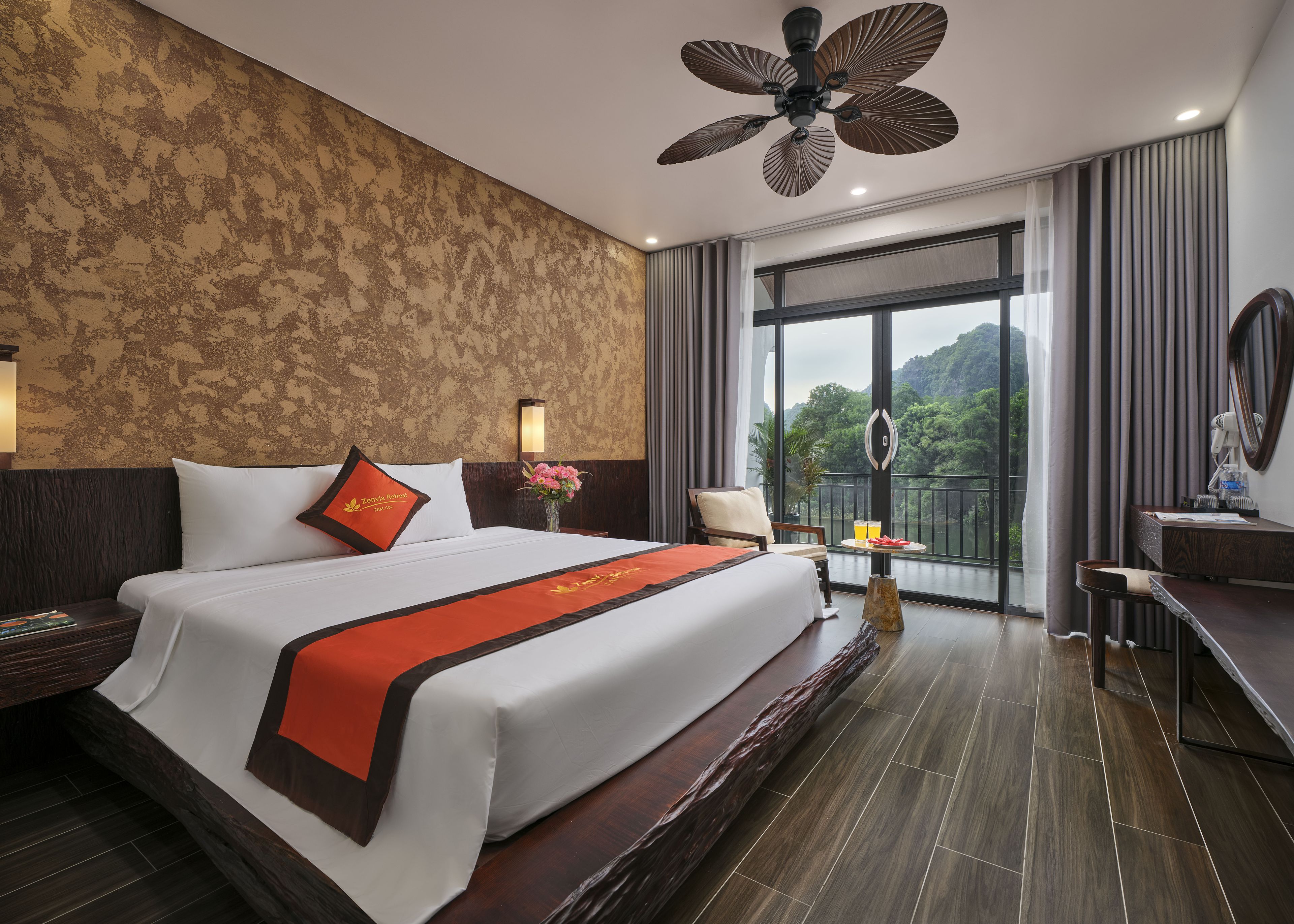 A hotel room with a large bed, a desk, a chair, a balcony with a view, and a ceiling fan.