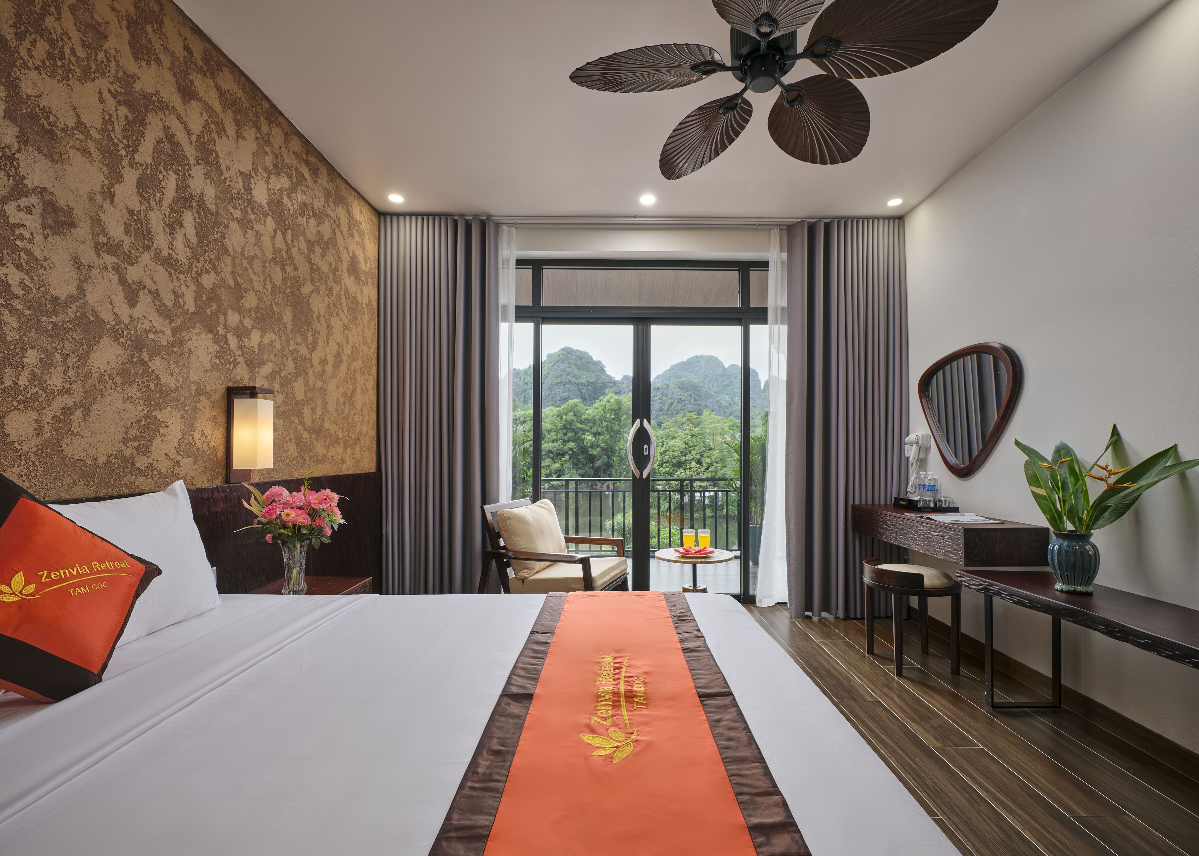 Ein Hotelzimmer mit einem großen Bett, einem Schreibtisch, einem Stuhl, einem Balkon mit Bergblick und einem Deckenventilator.