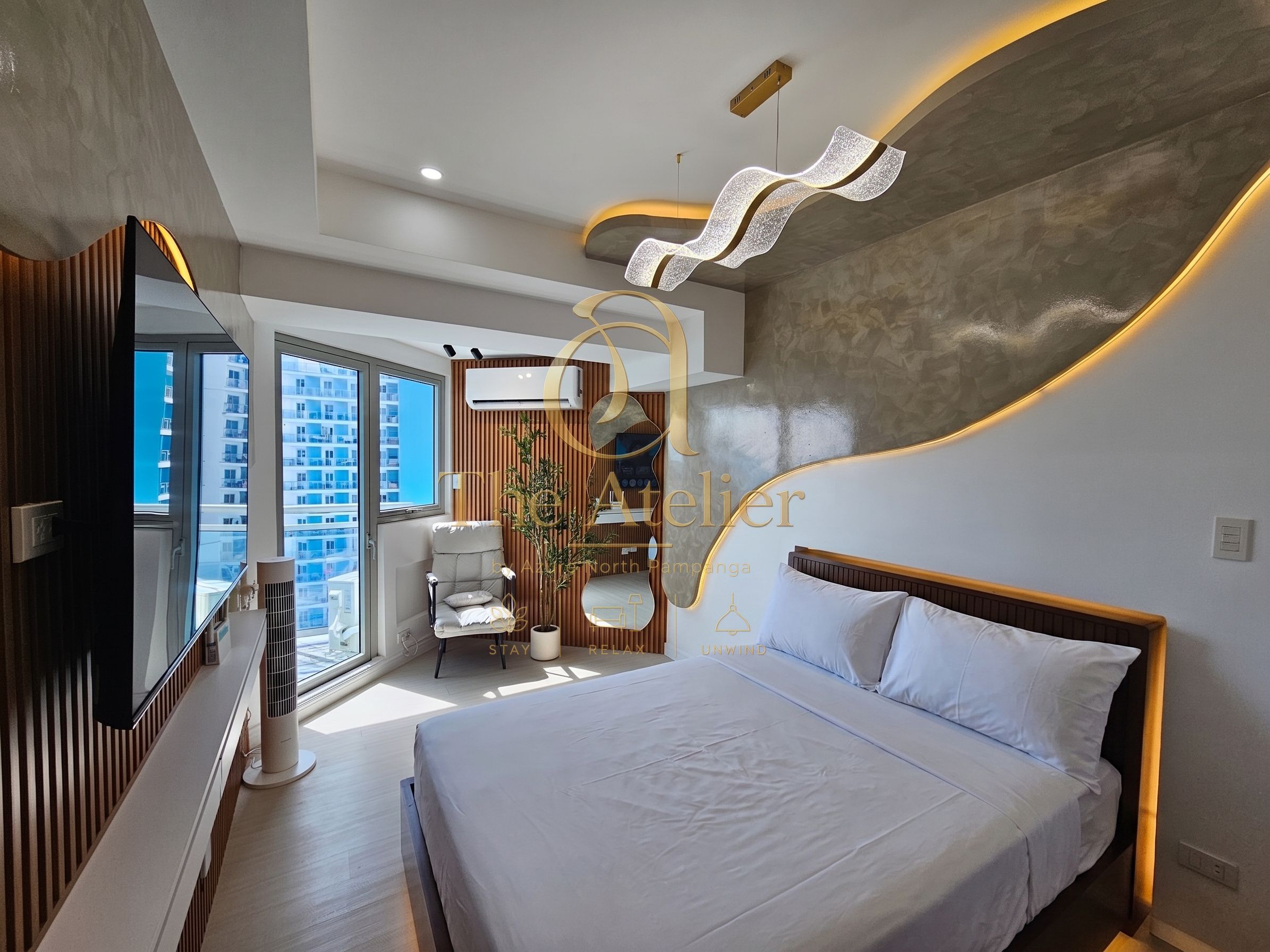 Une chambre d’hôtel moderne dotée d’un grand lit, d’un coin salon avec un fauteuil et offrant une vue sur la ville grâce à de grandes fenêtres.