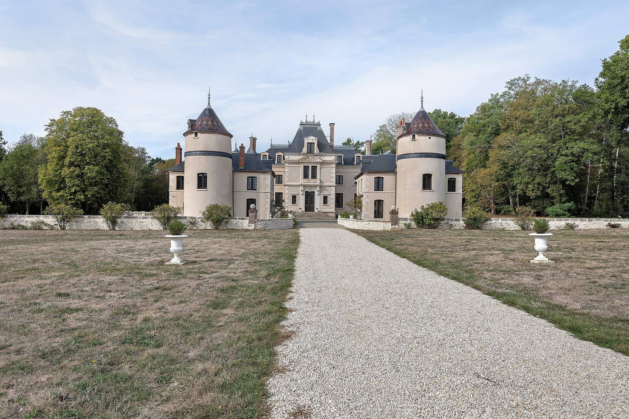 Un grand bâtiment symétrique, doté de nombreuses tours et d’une entrée centrale, entouré d’un chemin de gravier et de verdure.