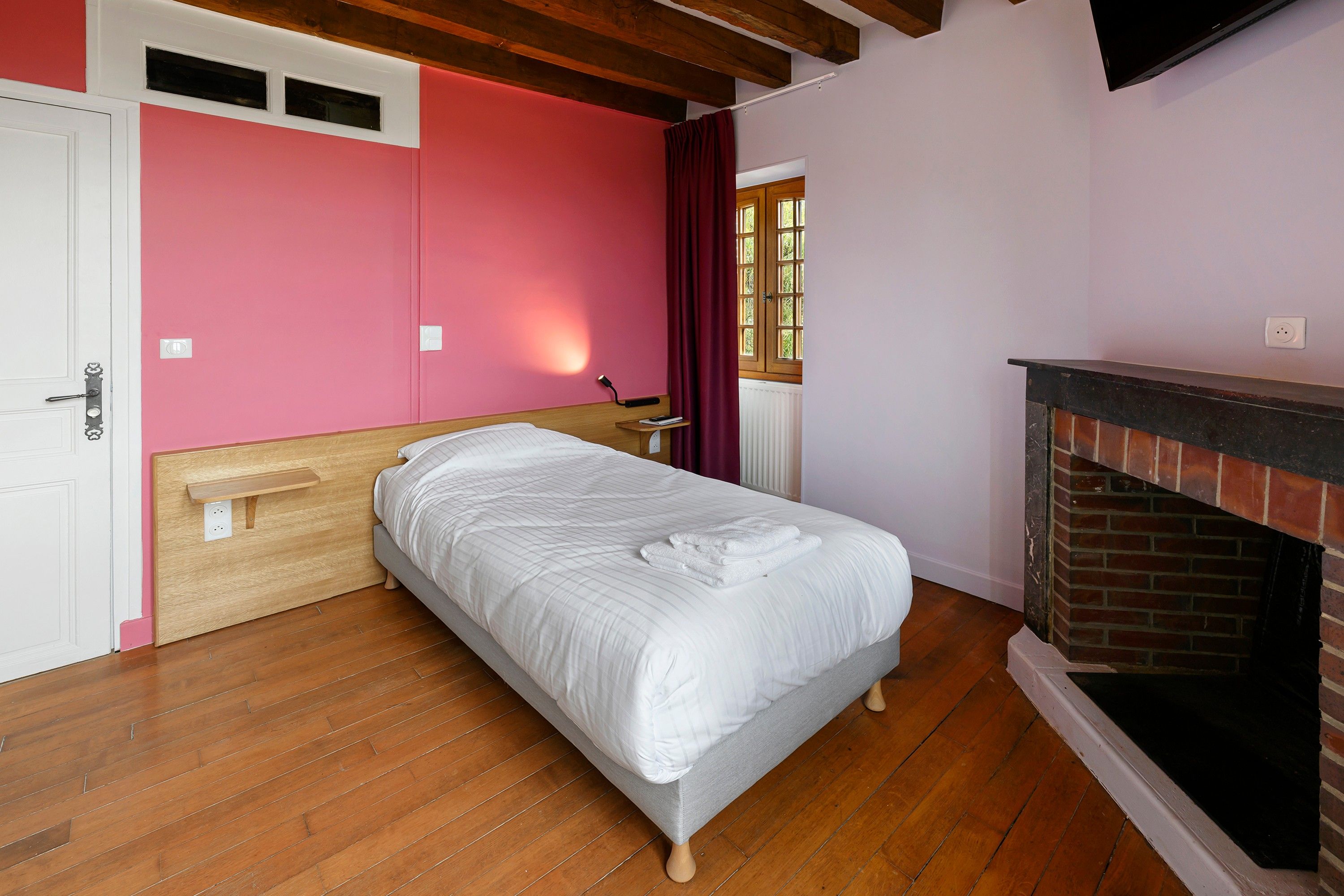 Een slaapkamer met een houten bed, een wit hoofdeinde, een bakstenen open haard en een raam met gordijnen.