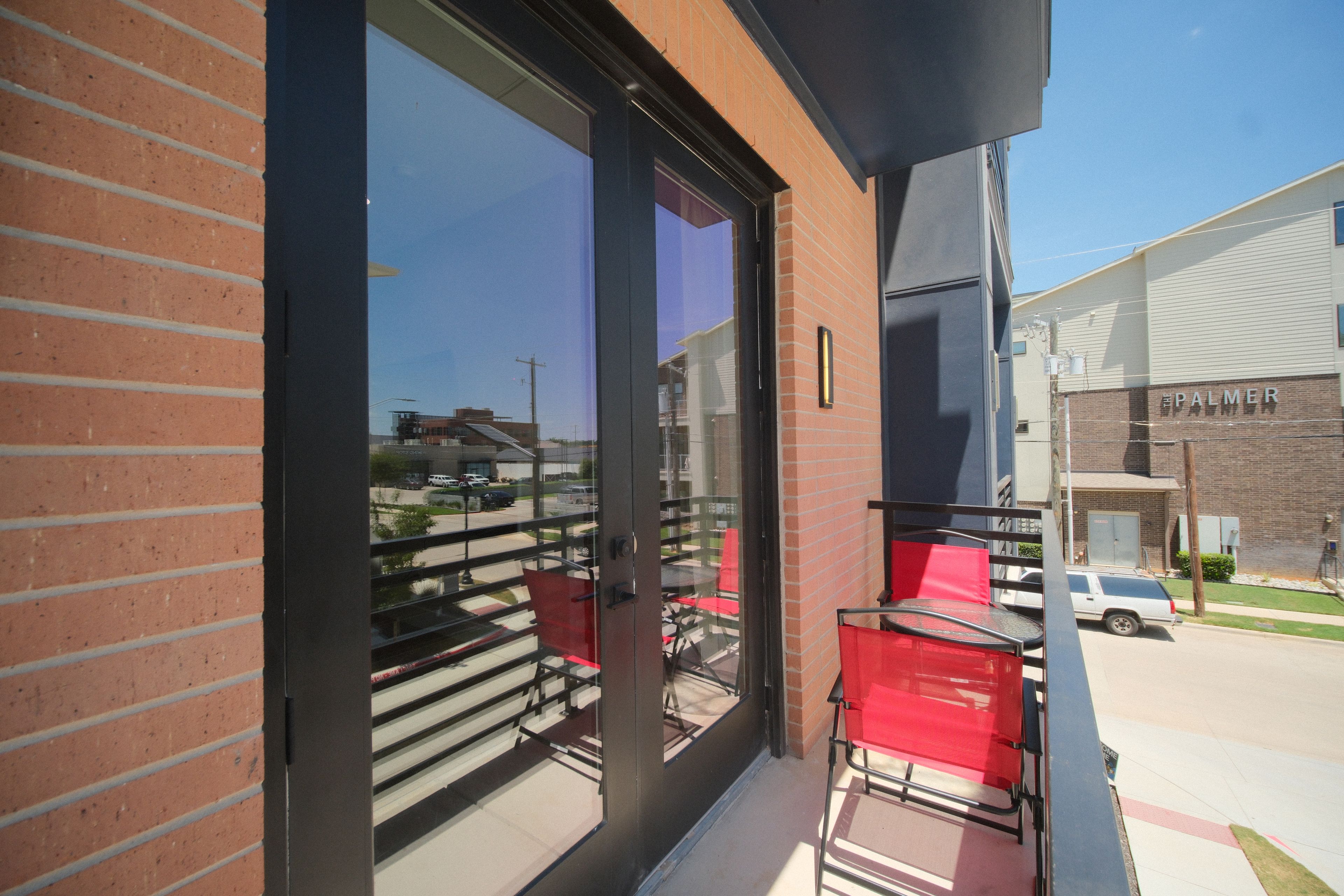 Un balcon avec une porte vitrée, des chaises rouges et une vue sur un parking et des immeubles.