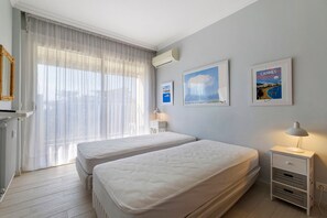 Ein Hotelzimmer mit zwei Betten, einem Fenster mit leichtem Vorhang, einer Wandklimaanlage und gerahmten Bildern.