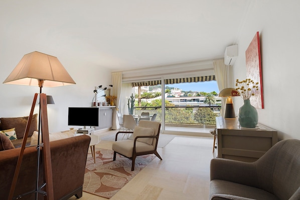 Ein Wohnzimmer mit Fernseher, Sofa, Sesseln, einer Lampe und Blick auf einen Balkon mit Pflanzen.