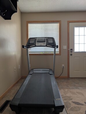 Un gimnasio en casa con cinta de correr, ventana con persianas y una puerta.
