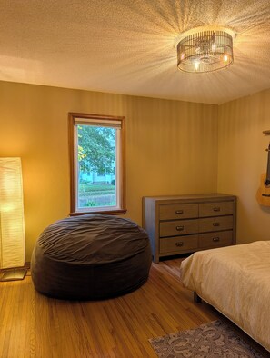 Un dormitorio con cama, cómoda, lámpara, ventana con vista a follaje y lámpara de techo.