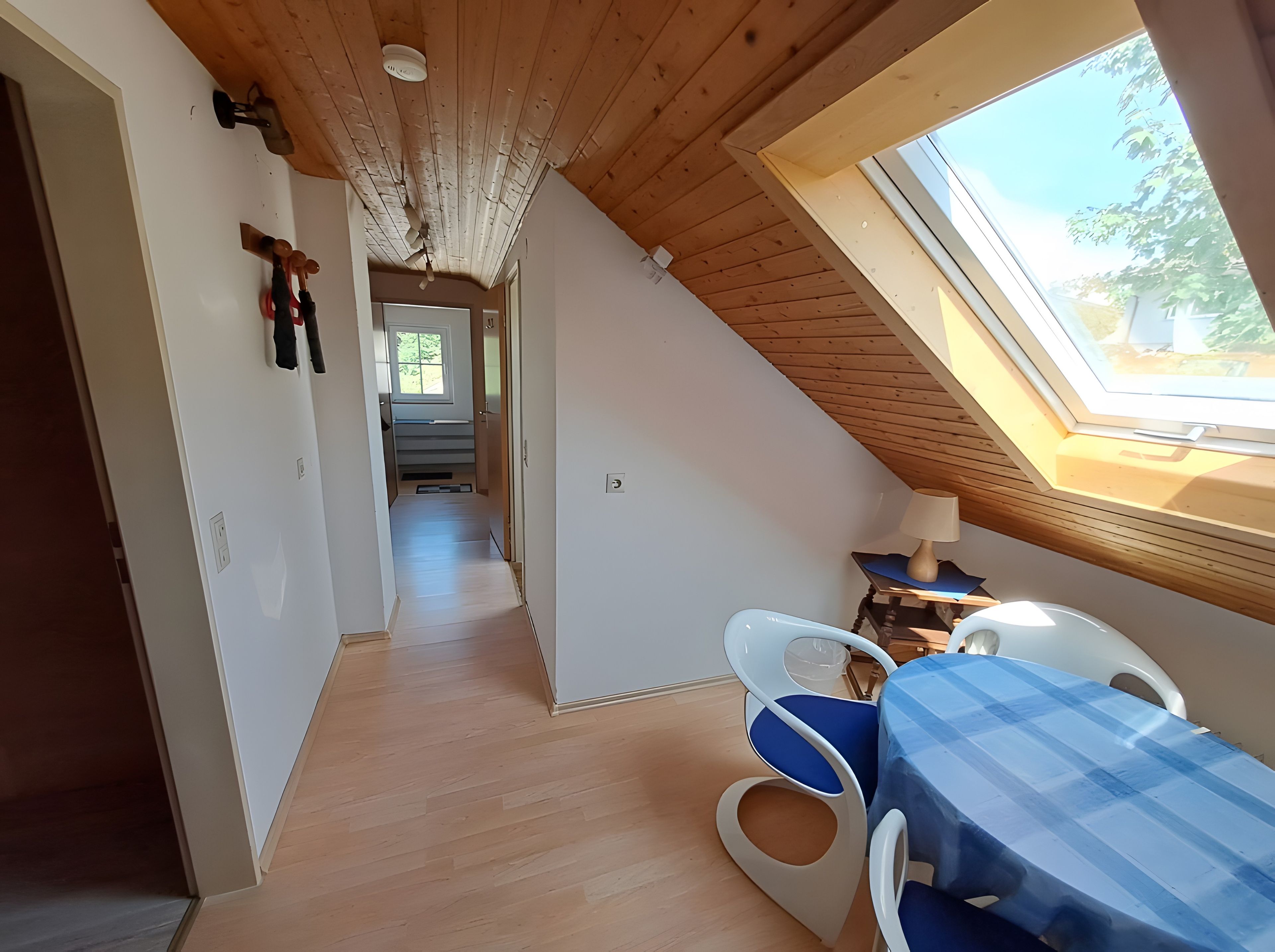 Ein Zimmer mit schrägem Dach, einem Oberlicht, einer Essecke mit Tisch und Stühlen sowie einem Gang zu einem weiteren Raum.
