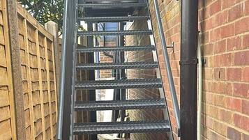 Escadaria de metal que leva à entrada de um edifício, cercada por um muro de tijolos e uma cerca de madeira.