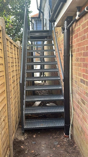 A metal staircase leading to a building entrance, surrounded by a brick wall and a wooden fence. - Apartment Sleeps 4 With Parking (Rye)