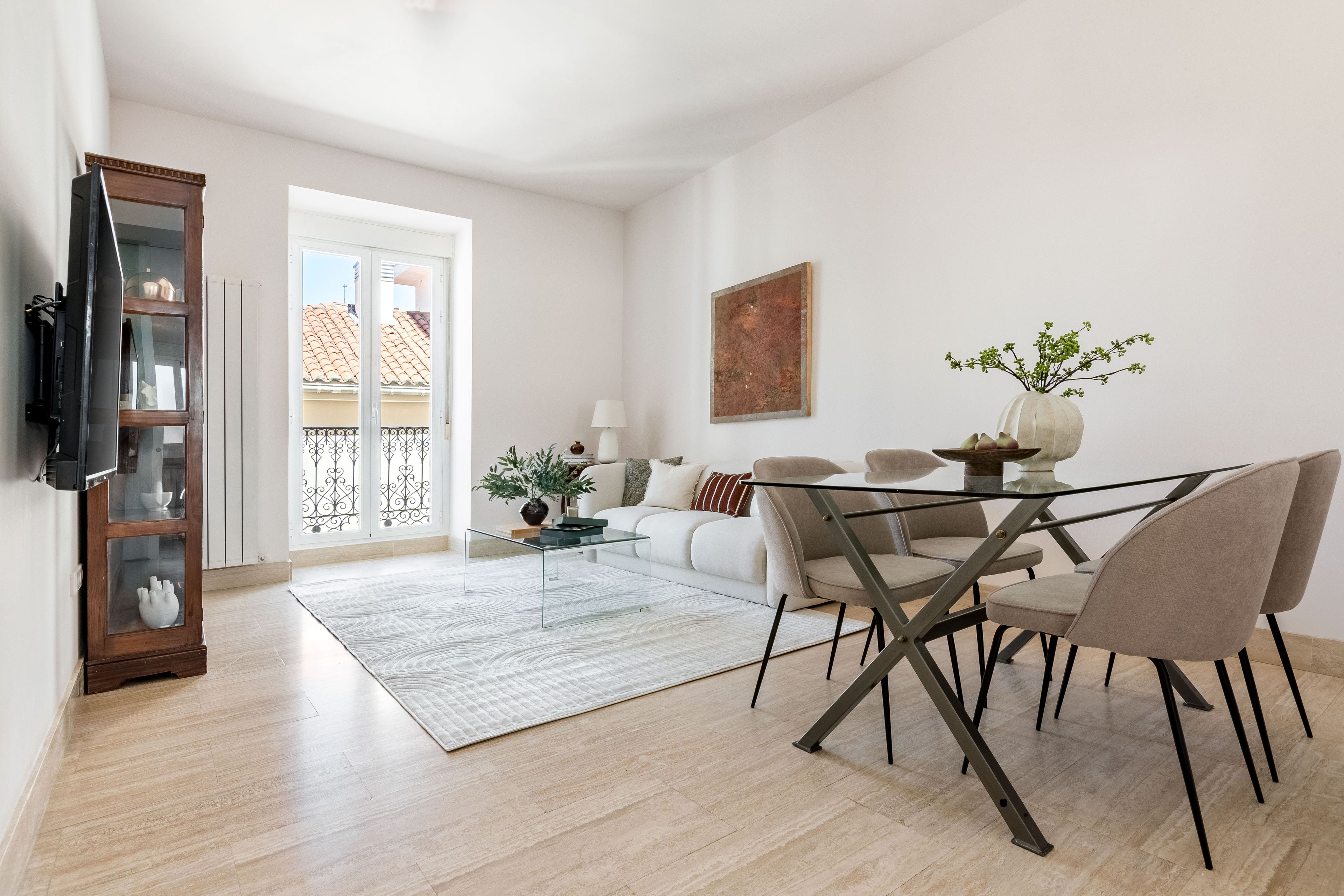 A modern living room with a glass coffee table, beige chairs, a white sofa, and a TV mounted on the wall.