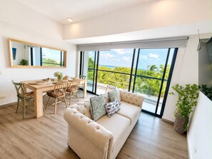 A modern living room with a sofa, dining table, chairs, and a large window with a view of greenery and the sky. - Luxurious 2BR Ocean View Condo Rooftop Pool and Jacuzzi (Cartagena de Indias)
