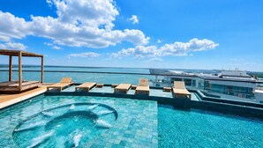 A rooftop infinity pool with wooden loungers and a hot tub, overlooking a body of water. - Luxurious 2BR Ocean View Condo Rooftop Pool and Jacuzzi (Cartagena de Indias)