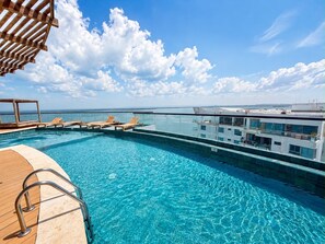 Une piscine à débordement située sur le toit, avec une terrasse en bois et des chaises longues, offrant une vue panoramique sur la ville.