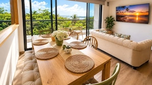 A modern living room with a large window overlooking a lush landscape, a dining area with a wooden table, and a sofa. - Luxurious 2BR Ocean View Condo Rooftop Pool and Jacuzzi (Cartagena de Indias)