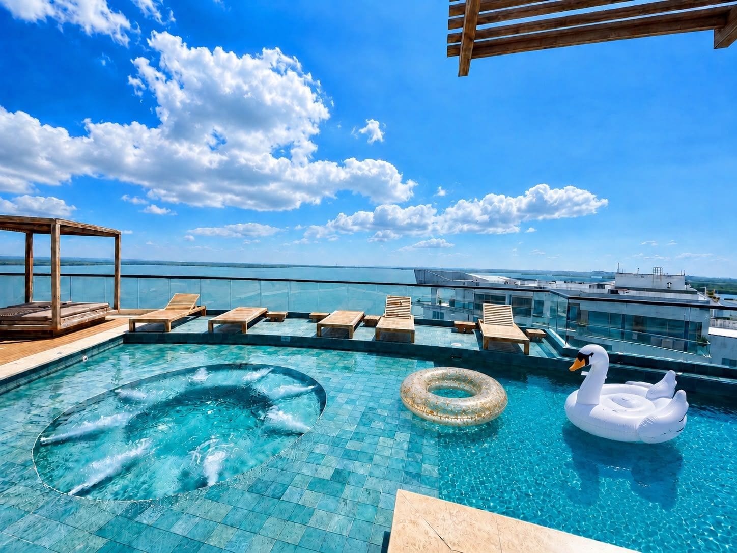 A rooftop infinity pool with lounge chairs and a swan float, overlooking a cityscape.