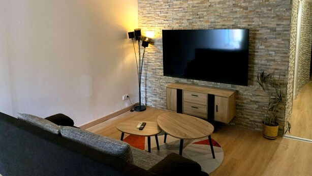 A living room with a flat-screen TV mounted on a stone wall, a sofa, a coffee table, and a floor lamp. - Le Cocon de Sohën – A Cozy and Comfortable Two-Bedroom Apartment in Montréjeau (MONTREJEAU)