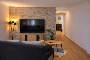 A living room with a stone wall, a flat-screen TV, a sofa, a coffee table, and a plant. - Le Cocon de Sohën – A cozy and comfortable two-bedroom apartment in Montréjeau (MONTREJEAU)