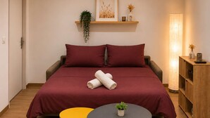 Un dormitorio con cama, mesita de noche, pequeña mesa redonda, una silla, una planta y estante con adornos.