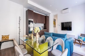 Ein moderner Wohnraum mit einem Esstisch aus Glas, einem blauen Sofa und einer Küchenzeile mit dunklen Holzschränken und Edelstahlgeräten.