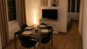 Un comedor con una mesa dispuesta para dos personas, un televisor sobre un mueble bajo y una cama visible a través de una puerta abierta.