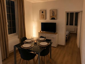 A dining area with a table set for two, a television on a stand, and a bed visible through an open door. - L'Escale d'Iris – A Cozy, Independent Two-Bedroom Apartment in Mazamet (Mazamet)