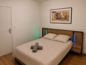 A neatly made bed with a patterned bedspread and pillows, a framed artwork on the wall, and a door with a handle. - L'Escale d'Iris – A Cozy, Independent Two-Bedroom Apartment in Mazamet (Mazamet)