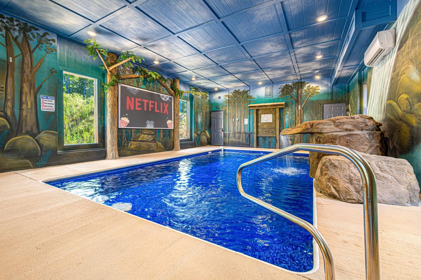 Piscina interna com sinal da Netflix, cercada por murais de árvores e pedras.