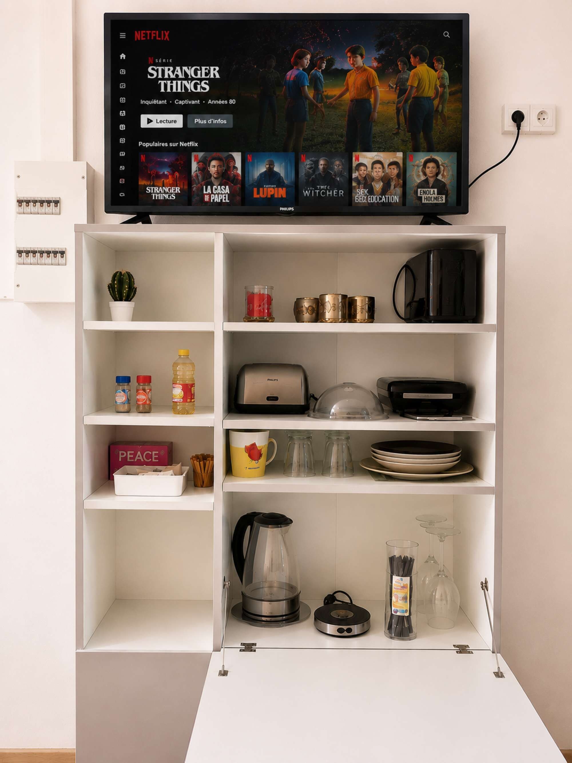 Une étagère blanche sur laquelle est posée une télévision diffusant la série « Stranger Things » de Netflix, ainsi qu’une sélection de livres et d’ustensiles de cuisine.