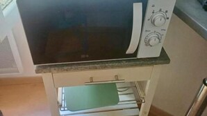 Eine Küche mit einer Mikrowelle auf einem weißen Standfuß, einem grünen Schneidebrett und einem Edelstahl-Wasserkocher.
