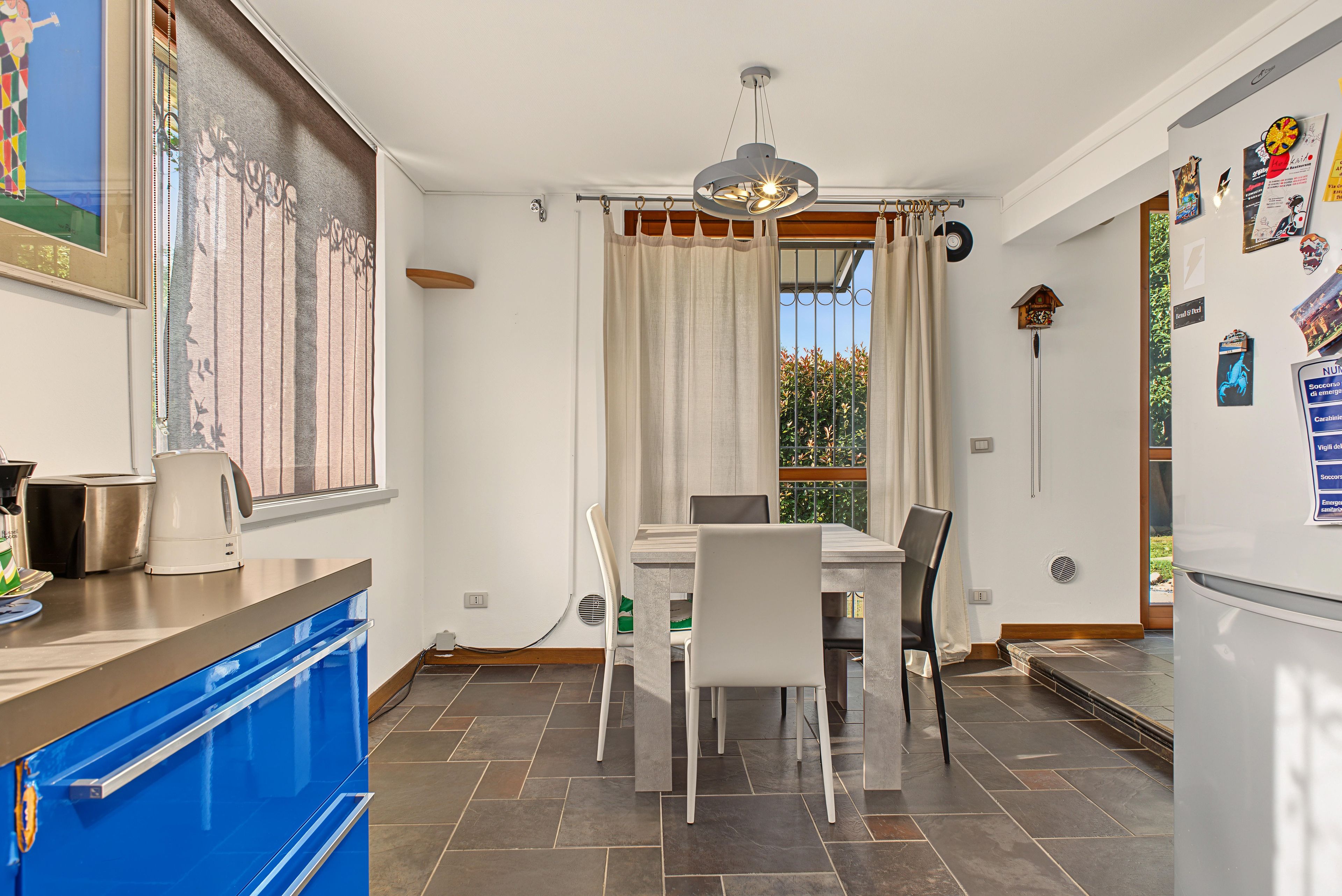 A modern kitchen with a dining area, featuring a window with curtains, a hanging light fixture, and a refrigerator with magnets.