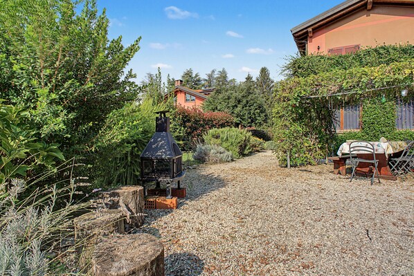A gravel patio with a barbecue grill, a wooden table with chairs, and a house in the background.