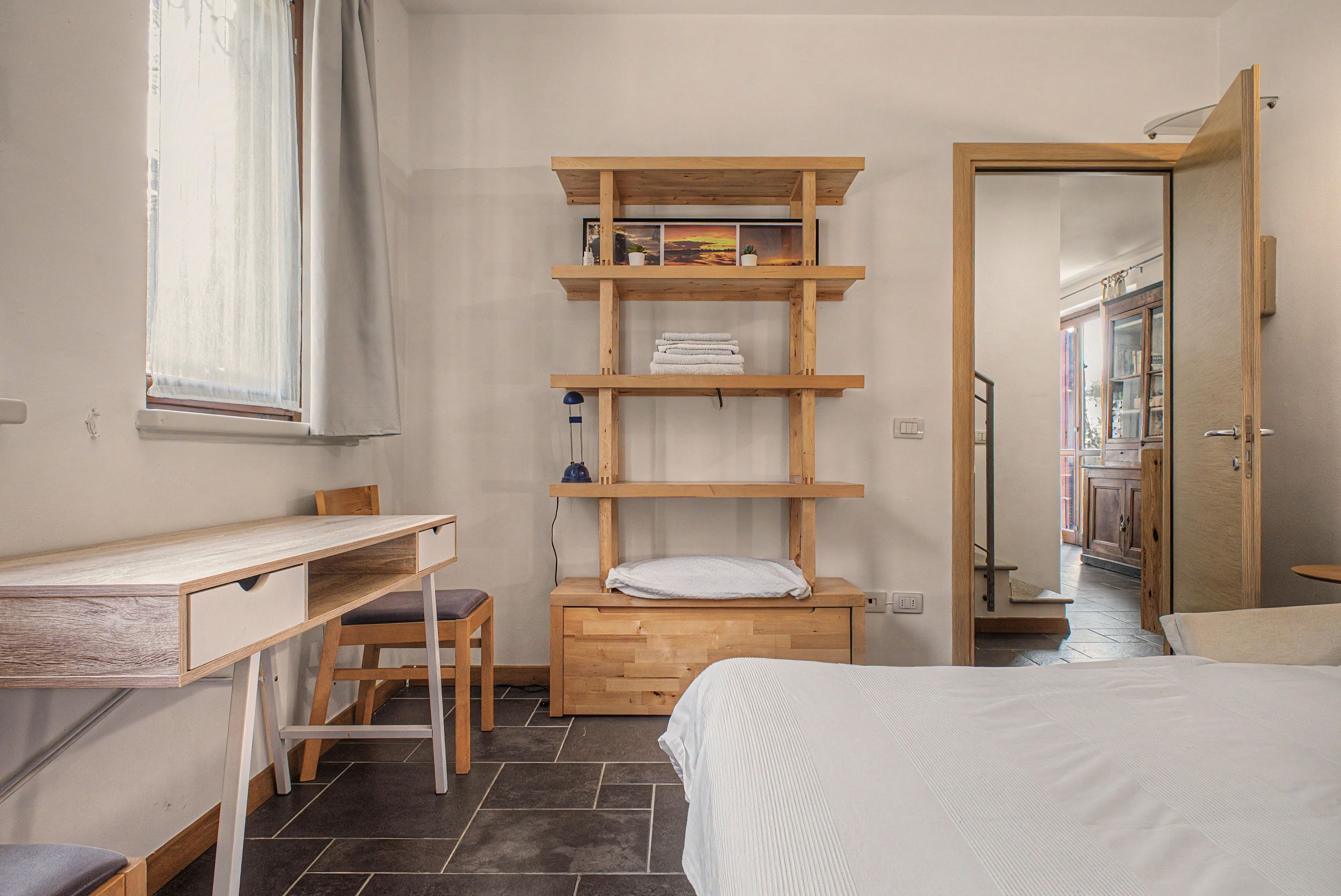 Un dormitorio con cama, escritorio, silla y estantería con toallas y una lámpara.