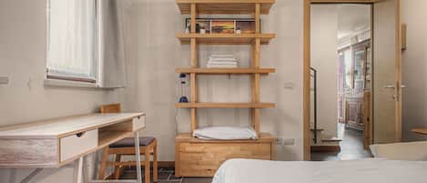 Un dormitorio con cama, escritorio, silla y estantería con toallas y una lámpara.
