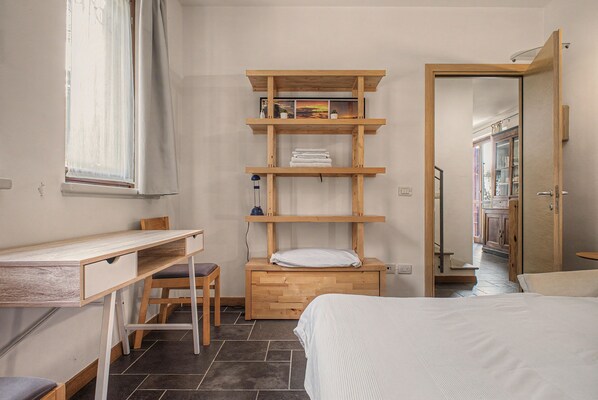 A bedroom with a bed, a desk, a chair, and a shelving unit with towels and a lamp.
