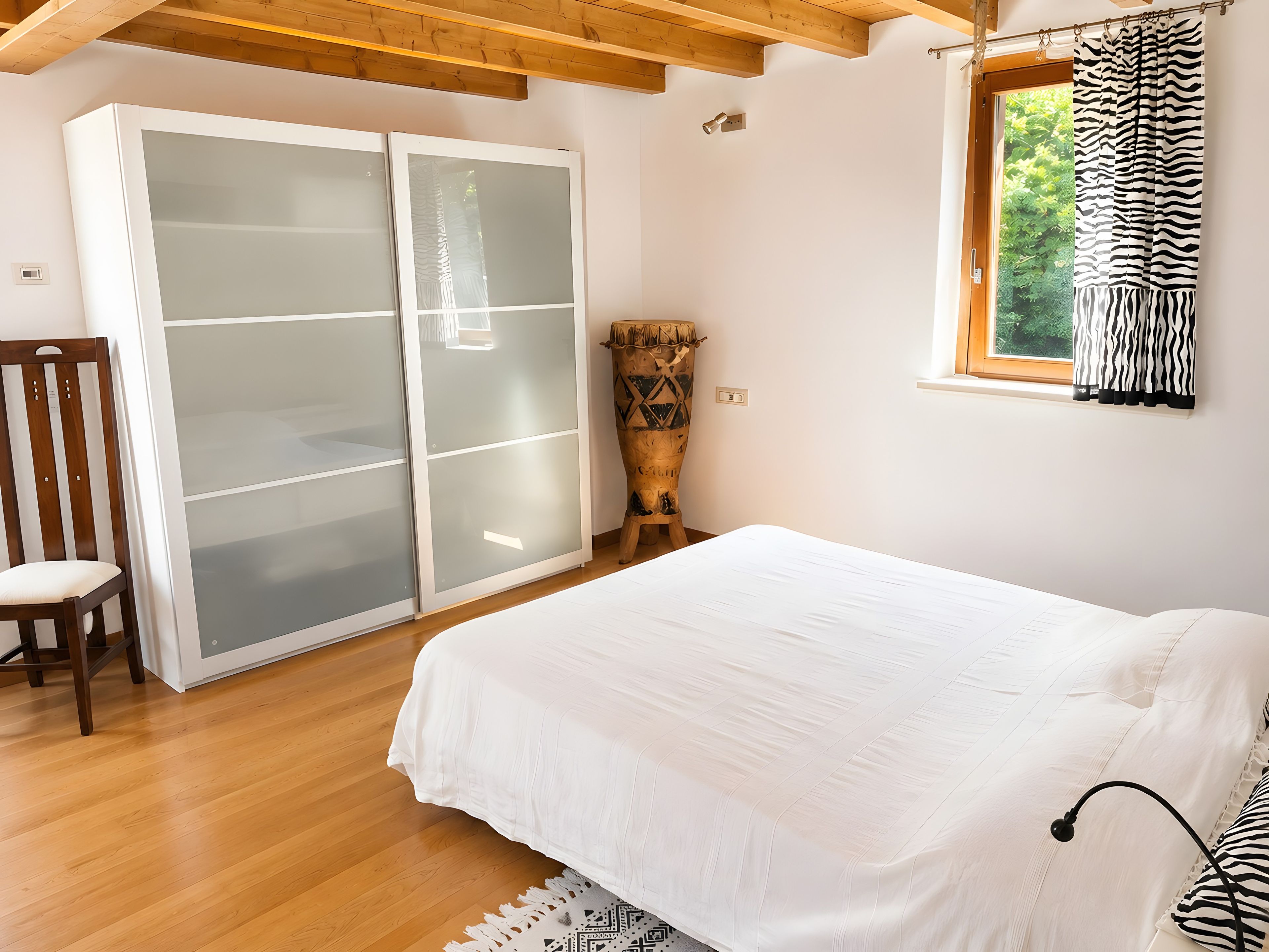 A bedroom with a bed, a wooden chair, a wardrobe with frosted glass doors, and a window with a zebra-striped curtain.