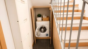 Un área de lavandería con una lavadora, un estante pequeño con una planta y botellas, y una escalera con barandilla.