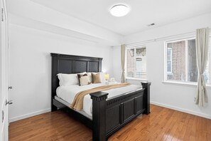 A bedroom with a wooden floor, a bed with a dark headboard, white walls, and a window with blinds. - A Foodie's Basecamp | 2BR/2BA Center City Philly (Philadelphia)
