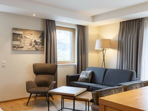 Ein modernes Wohnzimmer mit einer Couch, einem Sessel und einem Tisch. Es gibt ein Fenster mit Vorhängen, eine Lampe und ein Wandbild.
