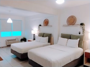 A hotel room with two beds, a bedside table with a lamp, a wall-mounted shelf with decorative items, and a window with blinds. - Suite 5 at Trinitaria House Hotel (San Juan)