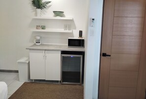 Eine kompakte Küchenzeile mit Mikrowelle, Kaffeemaschine und kleinem Kühlschrank. Weiße Schränke und eine Arbeitsplatte sind vorhanden.