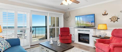 A living room with a television, red chairs, a fireplace, and a view of the ocean through the sliding glass doors.